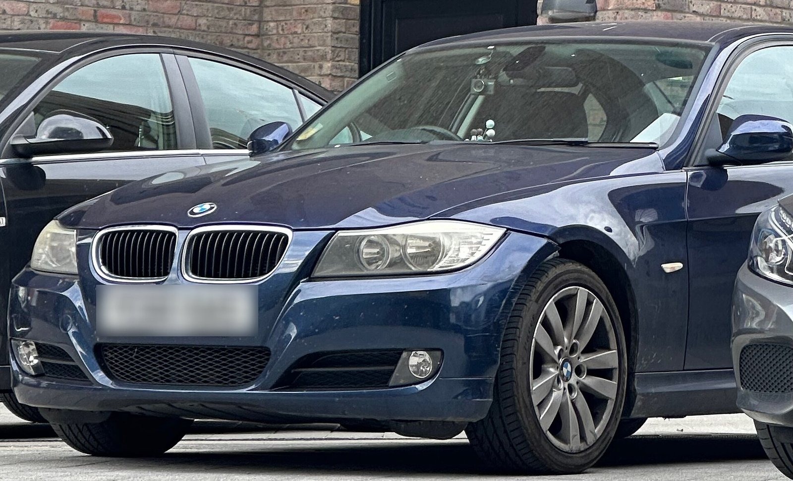 Zils BMW 3. sērijas sedans (E90) ar nieru formas restēm stāv uz ceļa, skats no priekšas trīs ceturtdaļās
